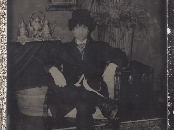 An old-time photo of a dapper gentleman in a bowler hat and suit sitting in a chair