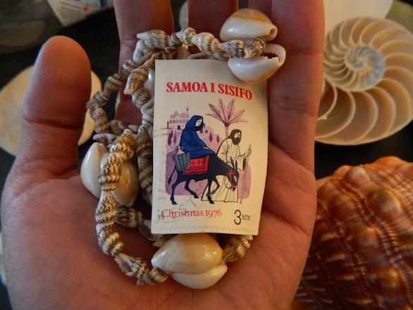 A hand holds a seashell necklace and a stamp from several decades ago in Samoa